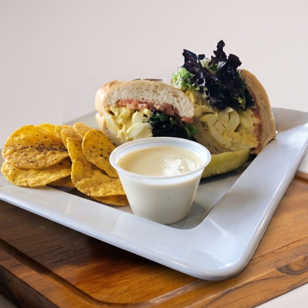 Egg salad sandwich with dark lettuce, tortilla chips, and a side of sauce on a white plate.