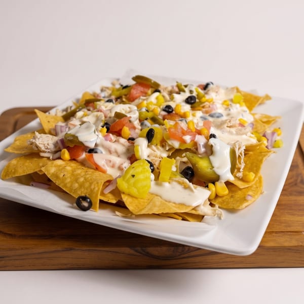 Loaded nachos topped with chicken, melted cheese, sour cream, black olives, corn, tomatoes, and jalapeños on a white plate.