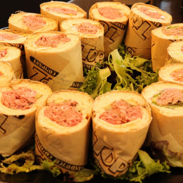Many rolled sandwiches, cut into sections, wrapped in "TOP SANDWICH COMPANY" paper on a platter with lettuce.