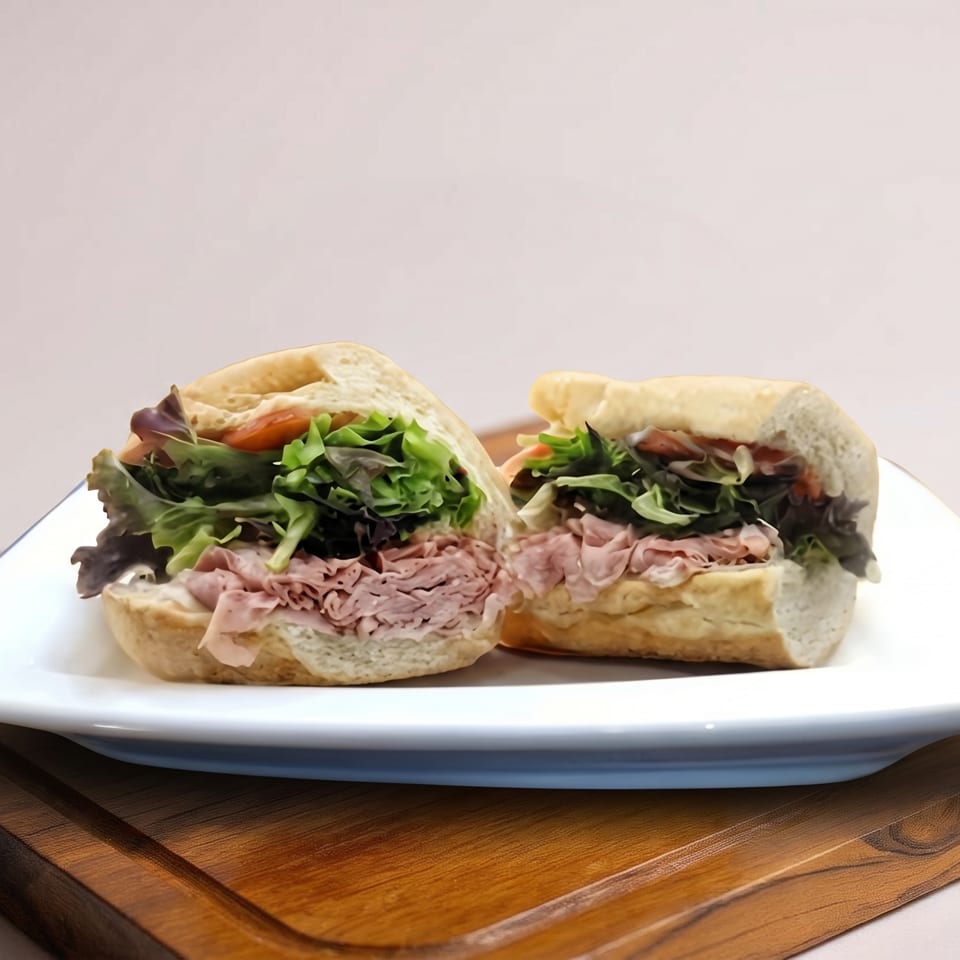 Two halves of a sandwich with meat and lettuce on a white plate over a wooden board.
