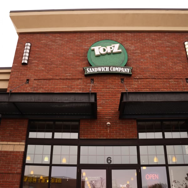 Brick-front Topz Sandwich Company restaurant entrance with green logo, black awnings, large windows, and illuminated open sign.