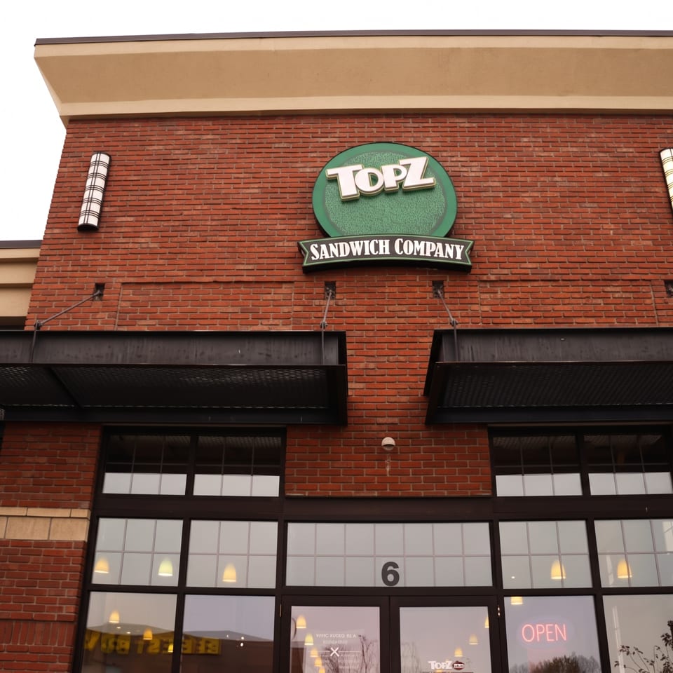 Brick storefront of Topz Sandwich Company featuring a green sign, black awnings, large windows, and an illuminated open sign.