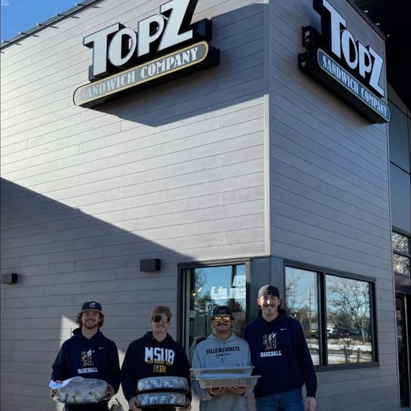 Four baseball players hold covered food trays outside a Topz Sandwich Company restaurant.