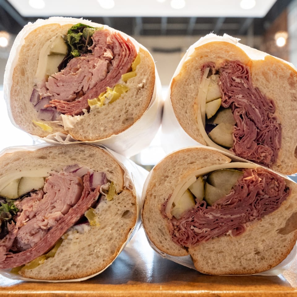 Four large deli sandwiches, cut in half, revealing layers of meat, cheese, and pickles.